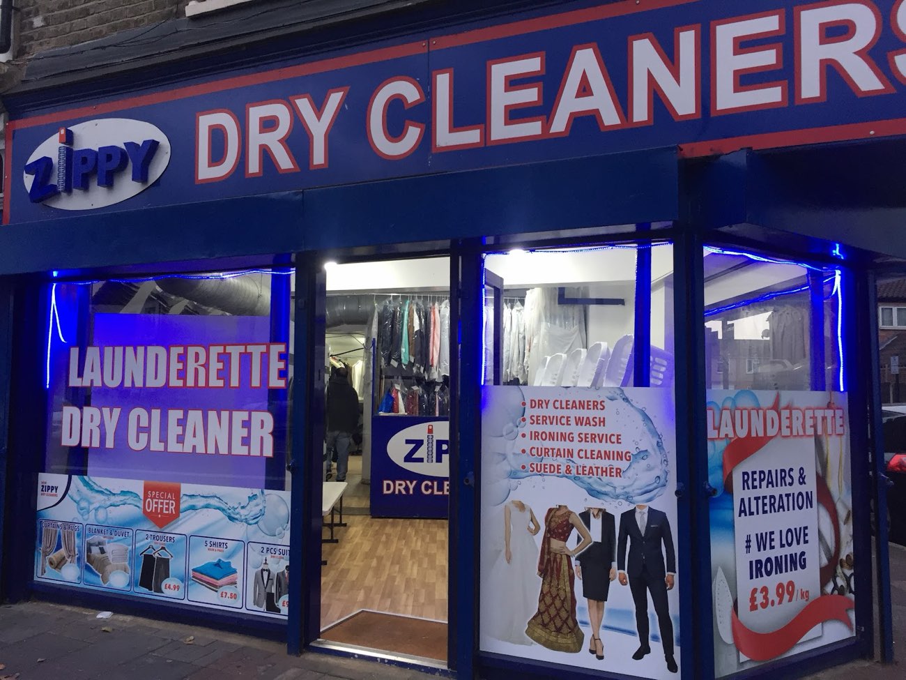 New Zippy Dry Cleaners shopfront on Barking Road lit up in the evening, garments visible on the rail inside